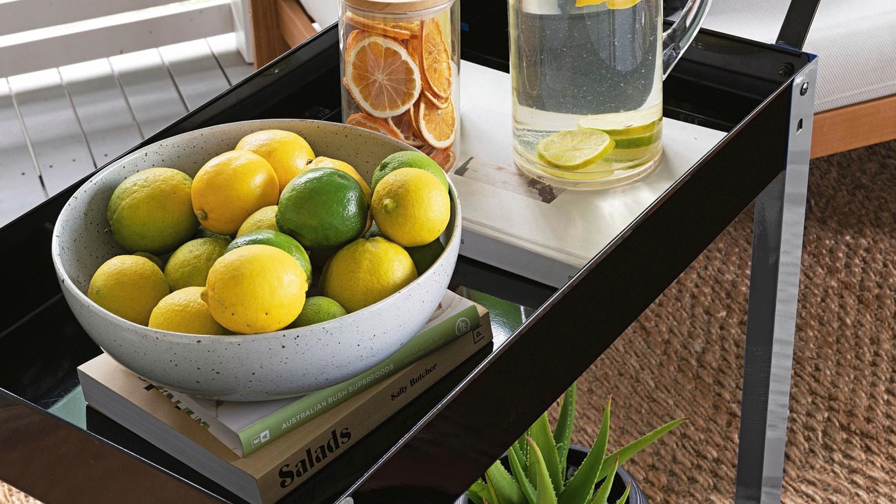 A black portable trolley on an outdoor deck in front of a lounge, with a bowl of fruit, a drinks station, books and plants.