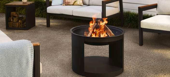 Black round outdoor fire pit with a visible flame, placed on a concrete patio and surrounded by modern outdoor lounge seating with light cushions and decorative pillows.
