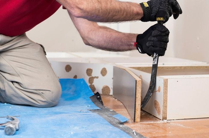 A person is using a pry bar to remove a kickboard from the base cabinet during a kitchen renovation.