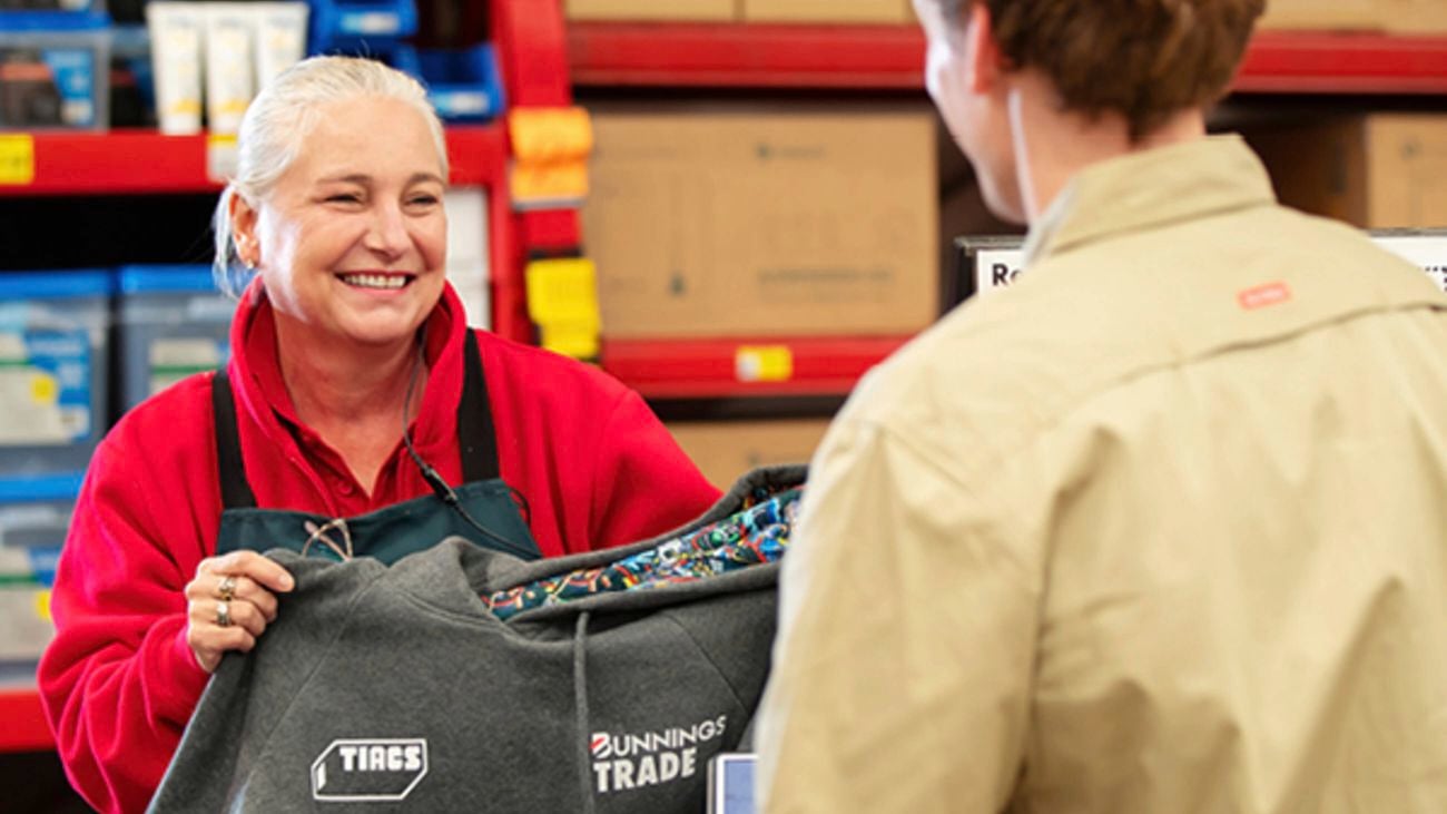 A Bunnings Team Member holding a TIACS grey hoodie and a customer standing in a Bunnings store at a counter.