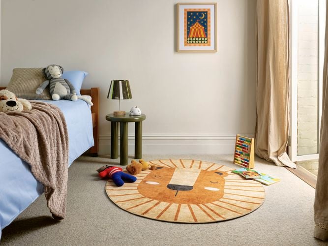 A child’s bedroom with a low bed, soft toys, a round lion-shaped rug, a small side table, and books near a sunlit glass door.