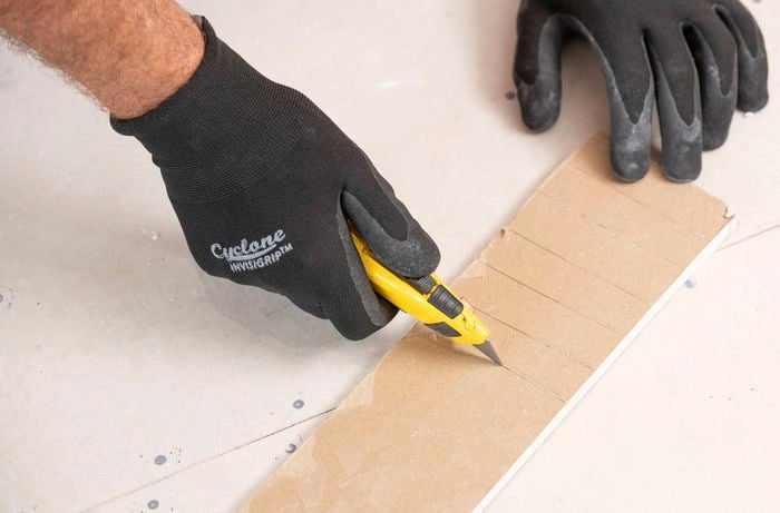 A person wearing black gloves is using a utility knife to score lines on the back of a plasterboard strip every 20 to 30 millimetres.