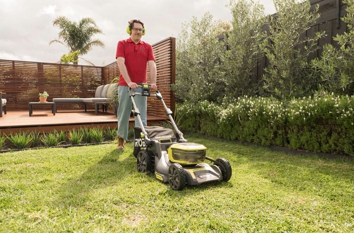 A Bunnings Team Member is mowing the lawn using a Ryobi electric lawn mower to prepare the lawn for topdressing
