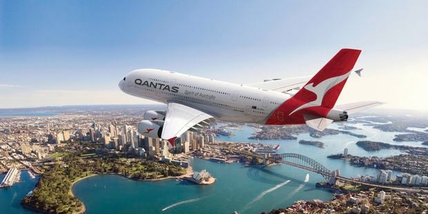 A Qantas plane flying over water and a city with a bridge across the water.