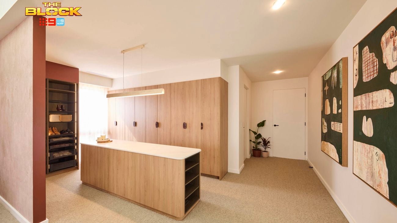A modern, minimalist room with a central wooden island topped with a white countertop, pendant lights above, and open shelving on one side. To the left is an open wardrobe with folded clothes and accessories. The back wall features built-in wooden cabinets with black handles. Two large abstract artworks in green and beige tones hang on the right wall. A potted plant sits near a door at the far end, and the floor is carpeted in a neutral tone.