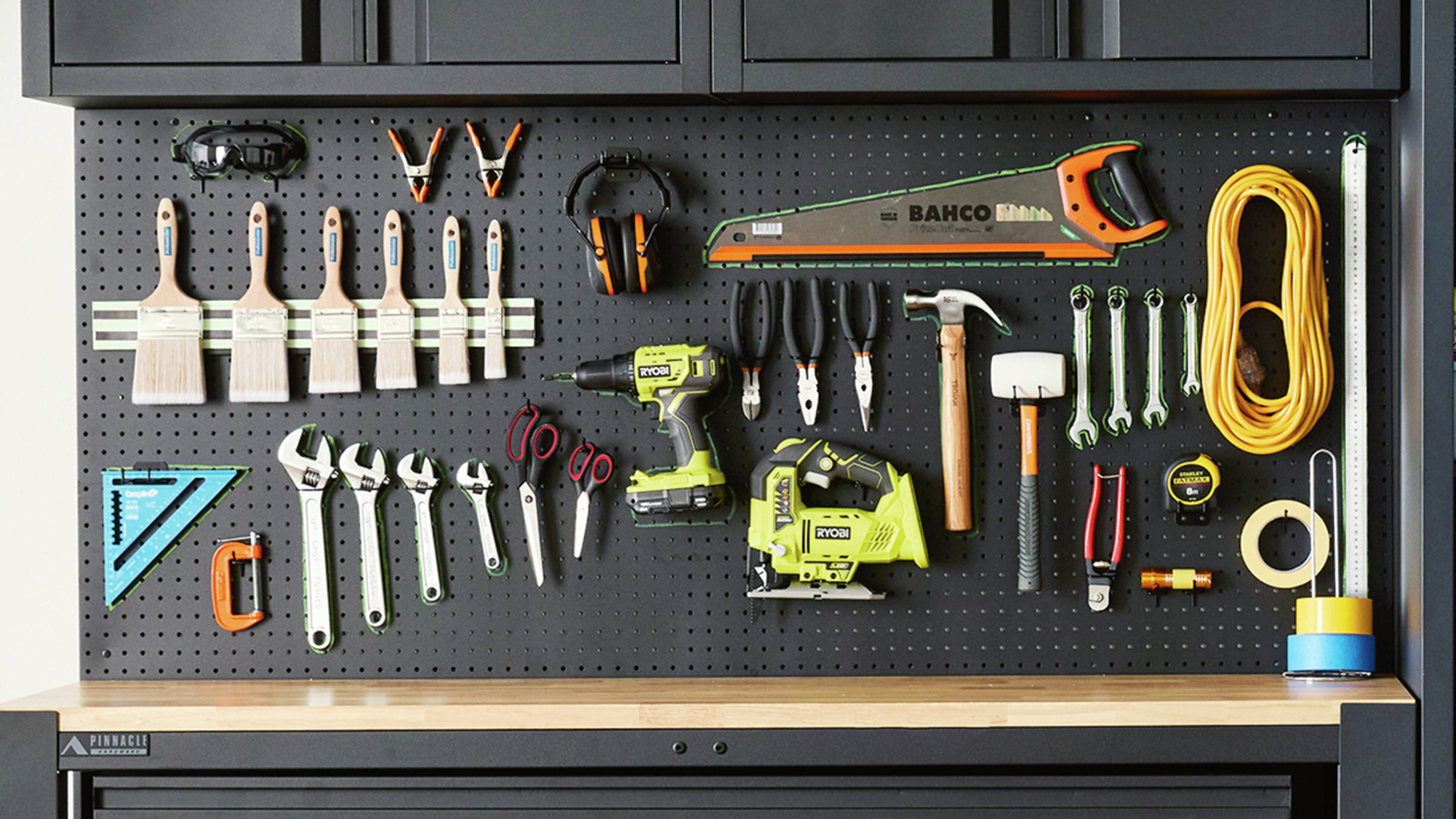 A workshop space with a black pegboard wall mounted over a timber workbench, featuring hand and power tools, plus safety equipment