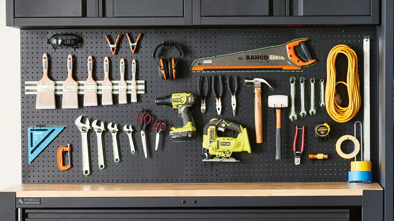 A workshop space with a black pegboard wall mounted over a timber workbench, featuring hand and power tools, plus safety equipment