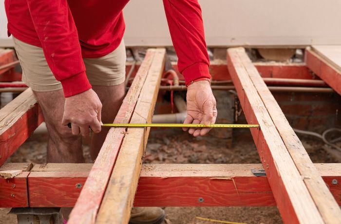 A person is measuring the gap between floor joists using a tape measure on a partially constructed wooden floor frame.   