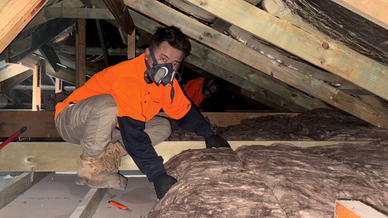 A man in orange hi-vis, wearing an aspirator mask and black gloves installs insulation