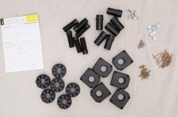 The image shows an organized layout of assembly hardware, plastic cabinet legs, screws, and an instruction sheet for a blind corner base cabinet.