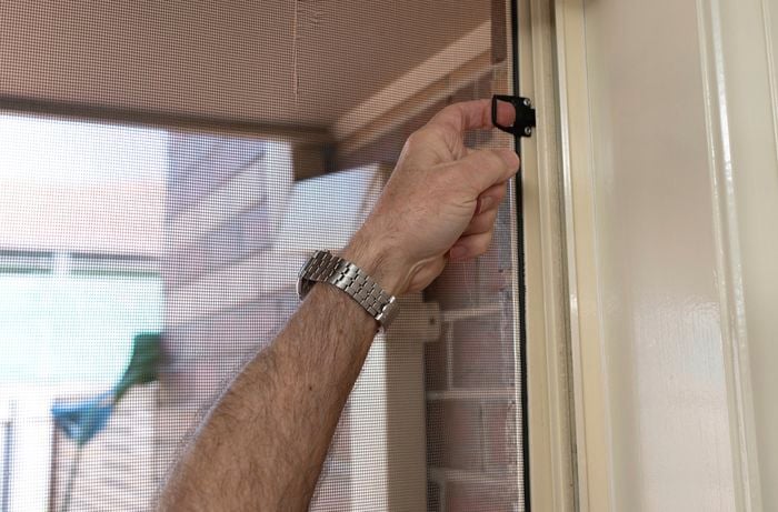 A person is removing a flyscreen from a window frame by releasing the clip that secures it.