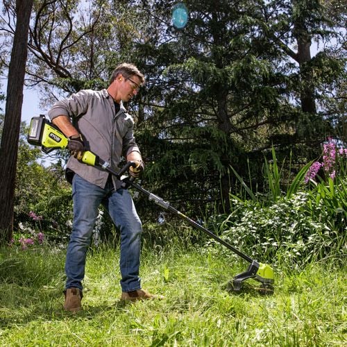 A person wearing protective gear and using a cordless line trimmer to cut the grass in a garden.