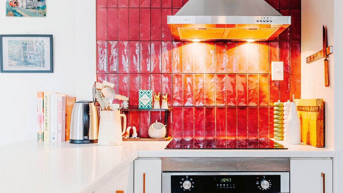 A kitchen with a red-tiled splashback, a stainless steel range hood, white cabinets with gold handles, and an oven in it.