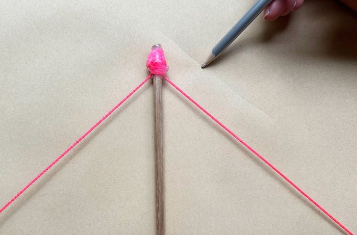 Vertical wooden dowel on brown paper with two neon pink strings taped to the top forming a V; a hand with a grey pencil points at the taped joint.