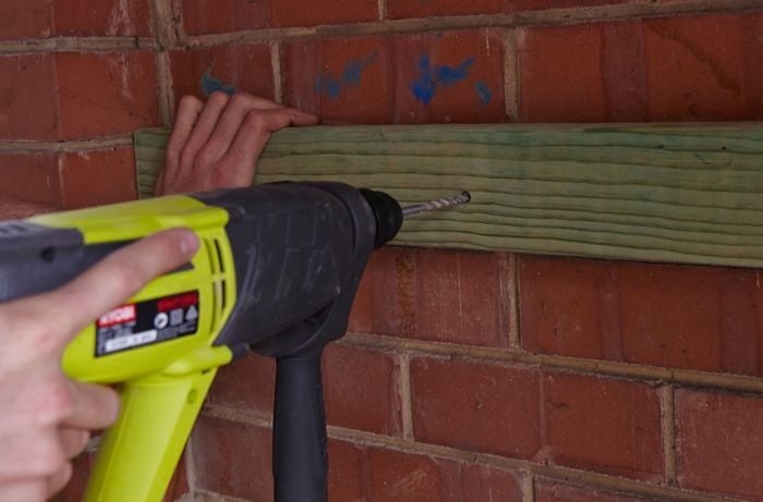 A person using a hammer drill to drill a hole a length of timber into a brick wall