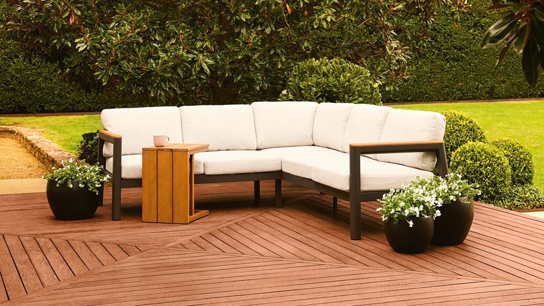 Outdoor corner lounge with a wooden side table on a timber deck, surrounded by green foliage.