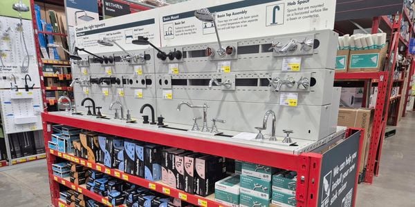 Tapware display in the plumbing section of a Bunnings Warehouse store, showcasing wall-mounted, basin-mounted and countertop tapware designs in chrome and matte black finishes. Educational signage explains tapware configurations, while shelves below are stocked with boxed tapware products and price labels.