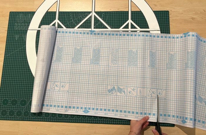 A left hand holds clear self-adhesive film which is being cut by scissors with a paper window template and green cutting mat underneath