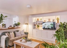 Wide view of a styled living room featuring a green sofa, coffee table, indoor plants, and white built‑in cabinetry with a television.