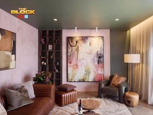 A modern living room featuring a large abstract painting on the wall, a brown leather sofa with cushions, an armchair, a wooden side table, and a floor lamp. A bookshelf with books and decorative items stands nearby. The room has soft lighting and curtains covering the windows. 