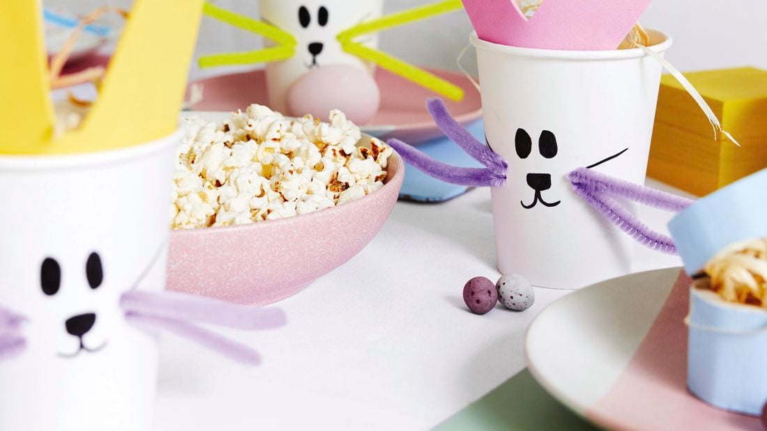 Craft cups with bunny faces on them next to a bowl of popcorn and decorative eggs.