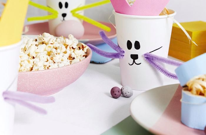Craft cups with bunny faces on them next to a bowl of popcorn and decorative eggs.