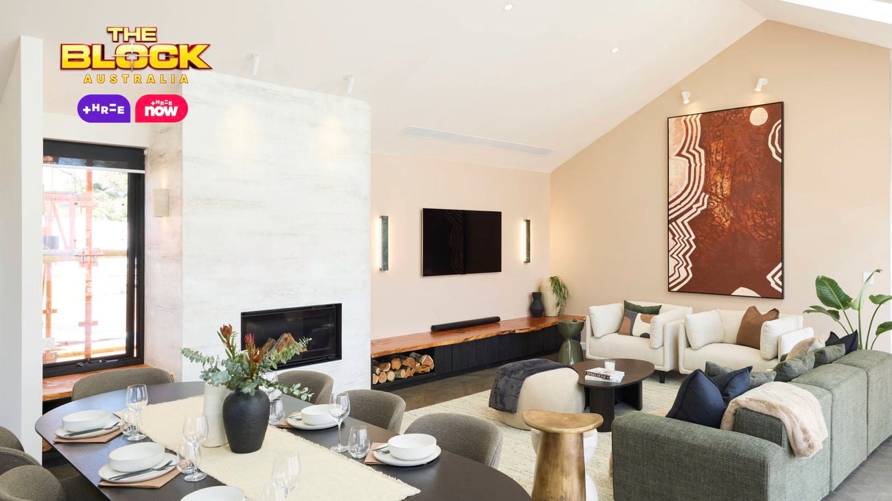 A modern open-plan living and dining area featuring a dining table set with plates, bowls, glasses, cutlery, and a floral centerpiece. The living room includes a large sectional sofa with cushions, a coffee table, and an abstract painting on the wall. A fireplace is built into a white marble wall, with firewood stored beneath a wall-mounted TV. 