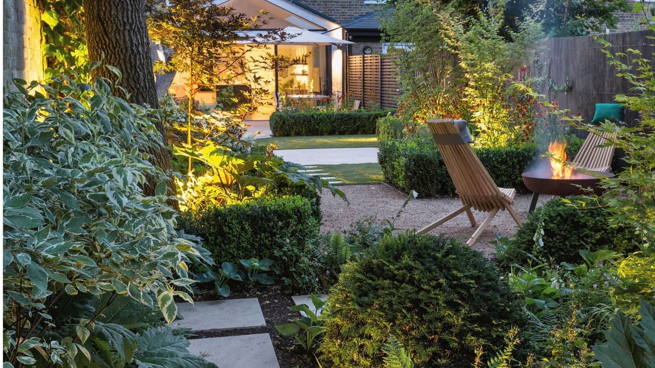 Garden with green shrubs surrounding two wooden outdoor chairs facing a lit fireplace