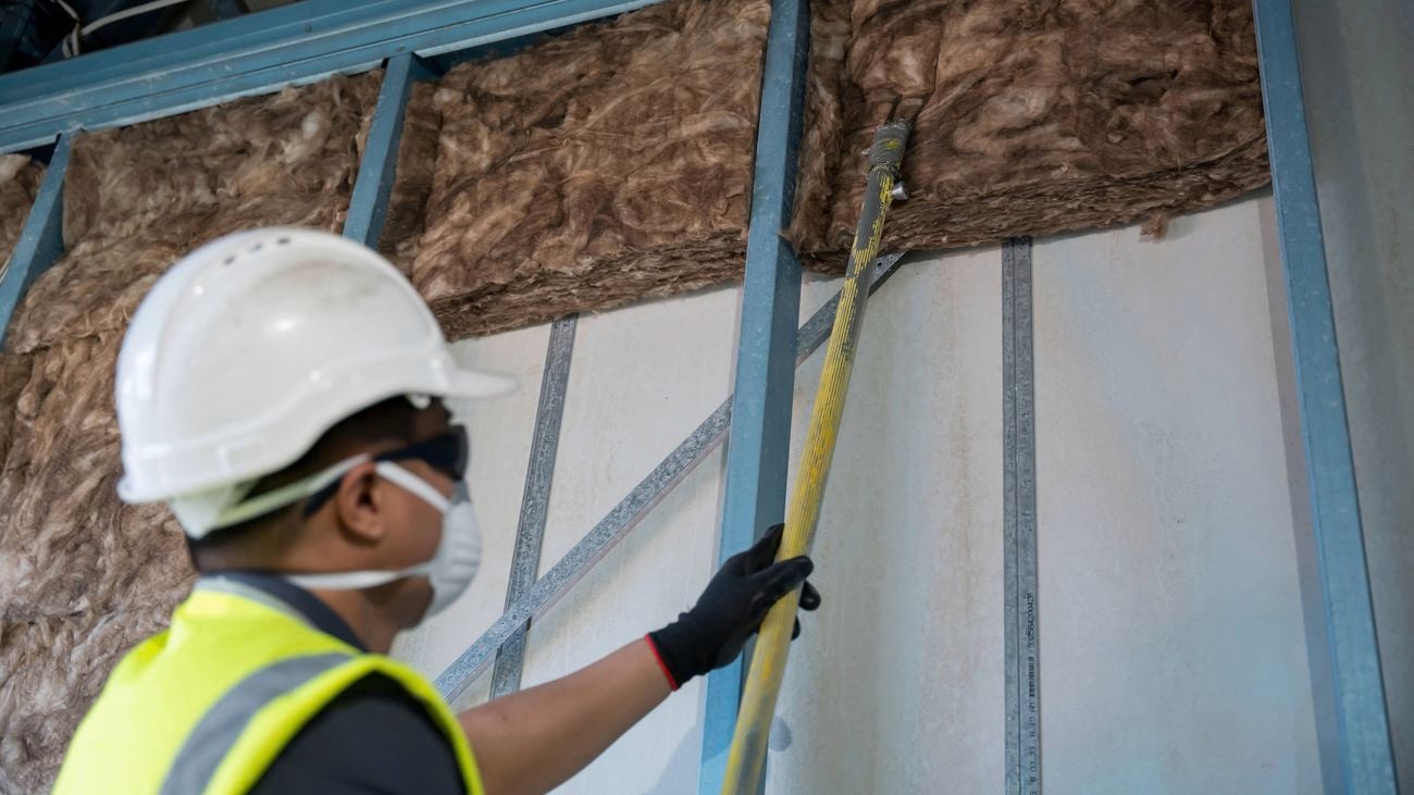 A worker in a hard hat, safety glasses, and face mask uses a long yellow tool to install fibrous insulation into a metal-framed wall cavity.]