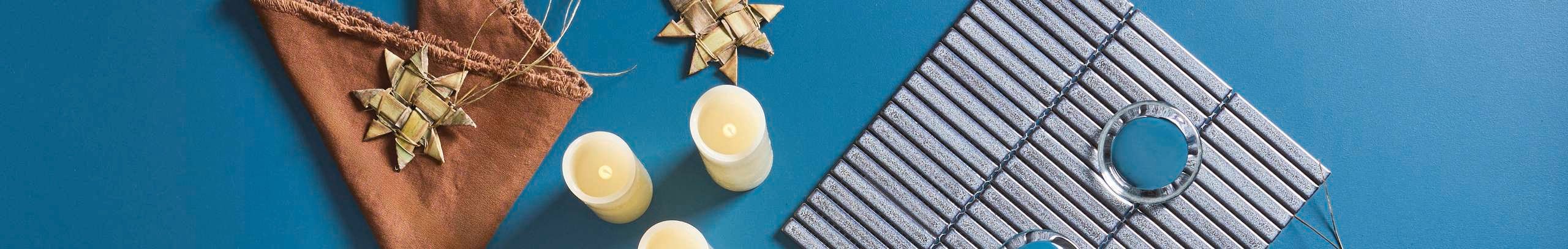 Flat-lay of a styled tabletop with woven star decorations, LED candles, a brown fringed cloth, and a tiled tray with glass holders on a teal background.