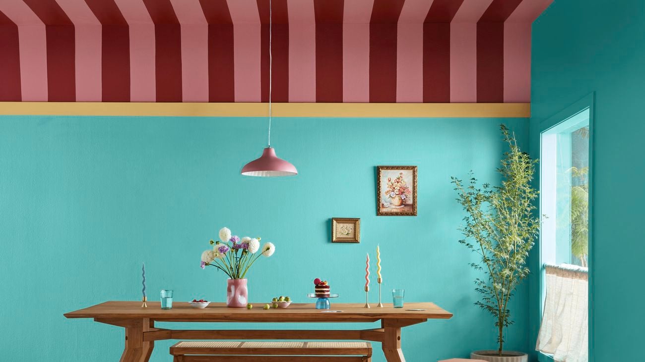 Dining room with blue walls and a red and pink striped ceiling. A tan dining table with seating benches sits in the centre of the room with a pot plant in the corner