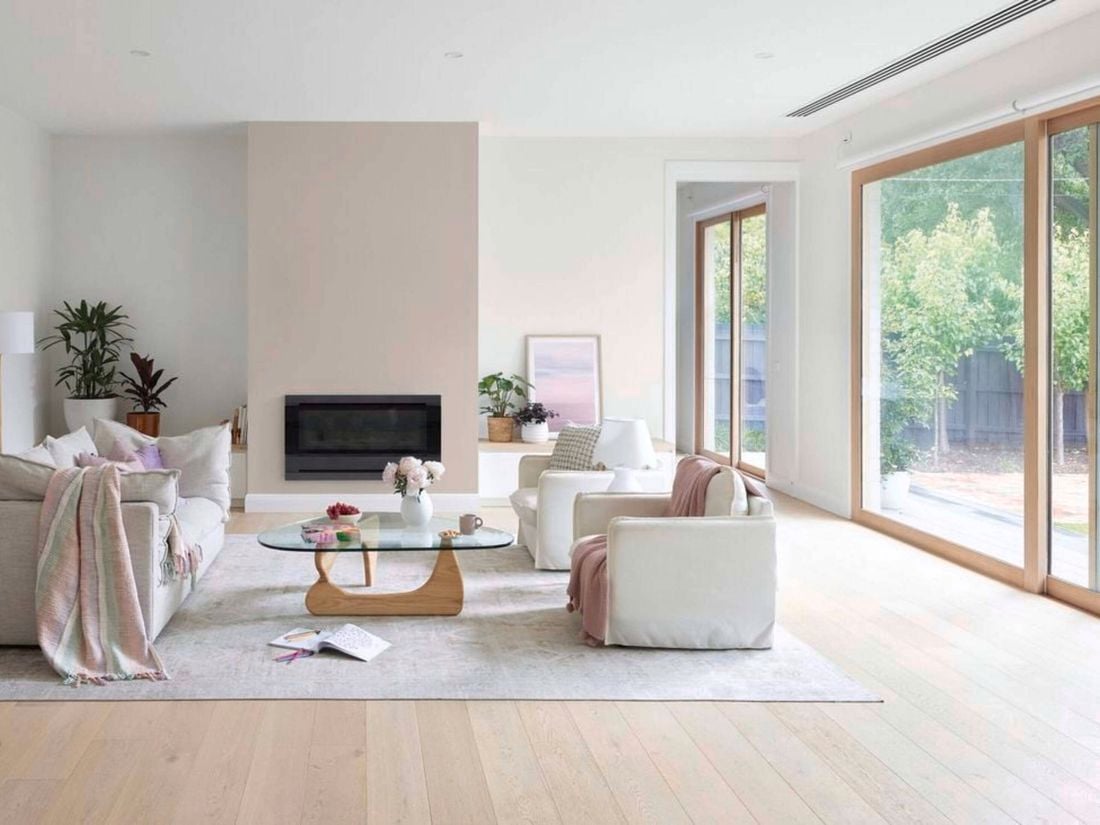 Bright, modern living room with light wood floors, neutral sofas, a glass coffee table, indoor plants, and large sliding glass doors opening to a garden.