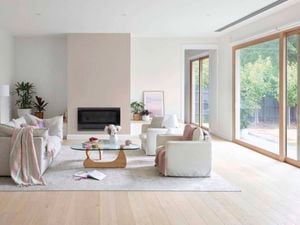 Bright, modern living room with light wood floors, neutral sofas, a glass coffee table, indoor plants, and large sliding glass doors opening to a garden.