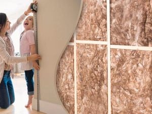 Woman measuring a childs height against a wall. To the right the wall is cut away to reveal insulation.