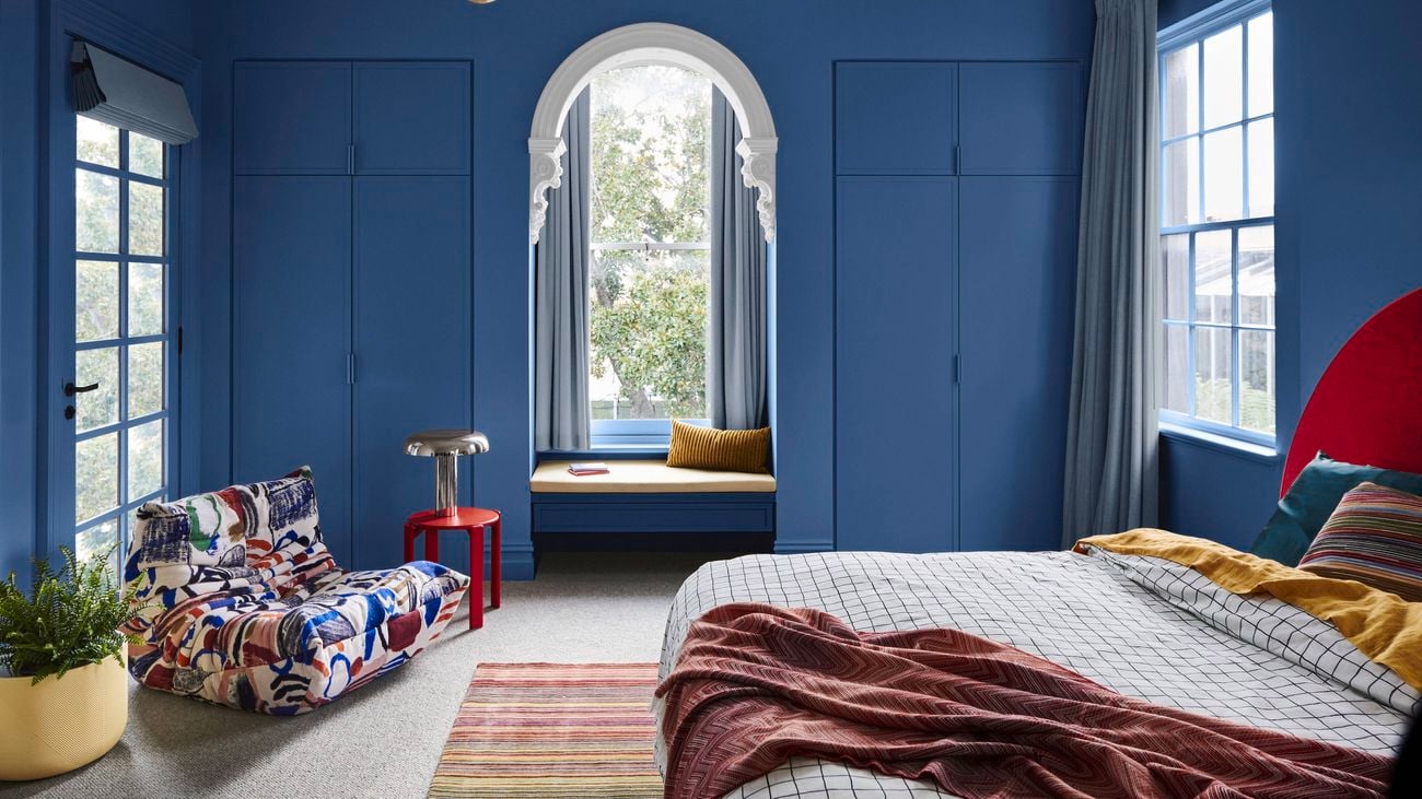 A bedroom with blue walls, a bed with a rug under it, a couch and a pot plant in it.