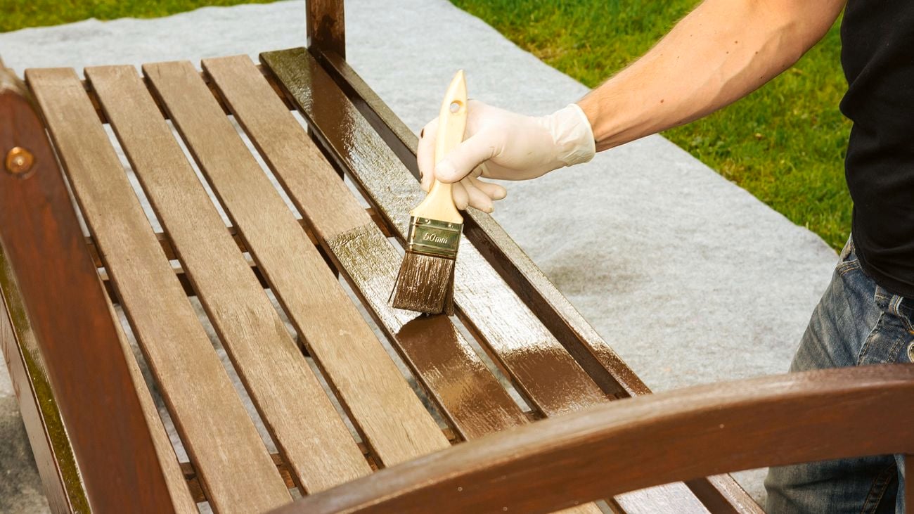A person wearing protective gloves uses a paint brush to oil an outdoor timber bench seat.