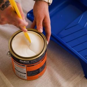 A hand with a tattoo is mixing Dulux paint using a brush, while a paint tray is kept on the side.