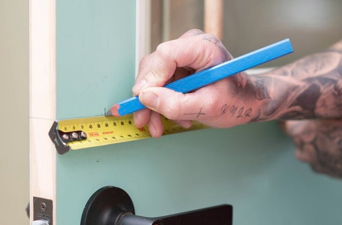 A hand is marking the backset on a door with a pencil while holding a tape measure. 