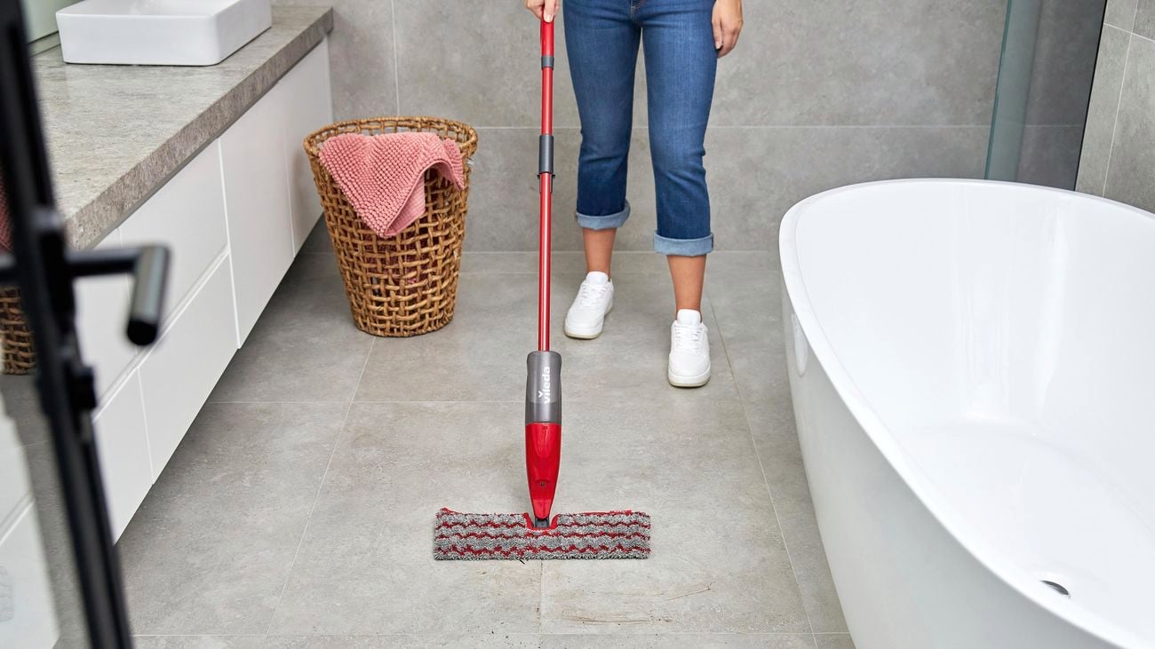 A woman wearing white sneakers uses a Vileda spray mop to clean grey tiles