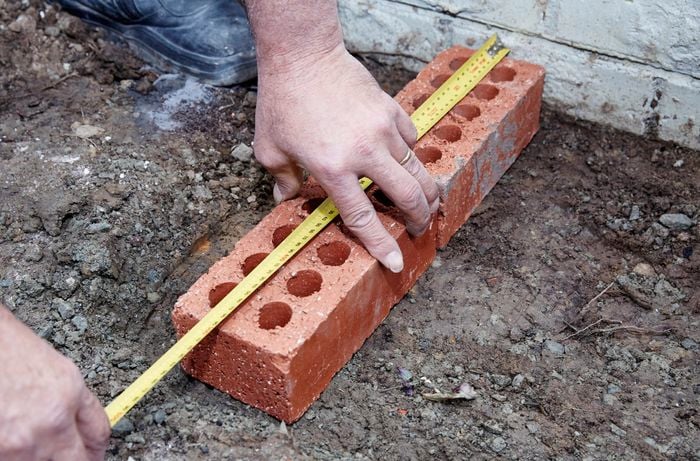 A person measuring two bricks laid at right angles to a house wall