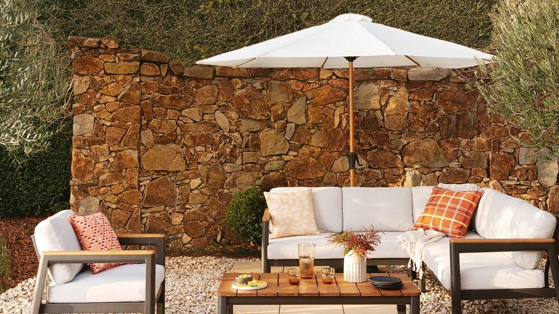 Outdoor patio lounge setting featuring a white-cushioned sectional sofa and matching armchair around a wooden coffee table on white gravel. A large white umbrella shades the seating area, with a rustic stone wall, manicured hedge, and olive trees creating a landscaped garden backdrop. Decorative cushions, a vase of flowers, and drinks on the table add a warm, inviting atmosphere.