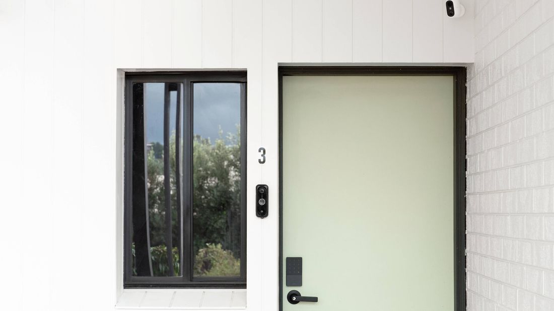 Front door with smart lock, video doorbell and security camera on a white house exterior.