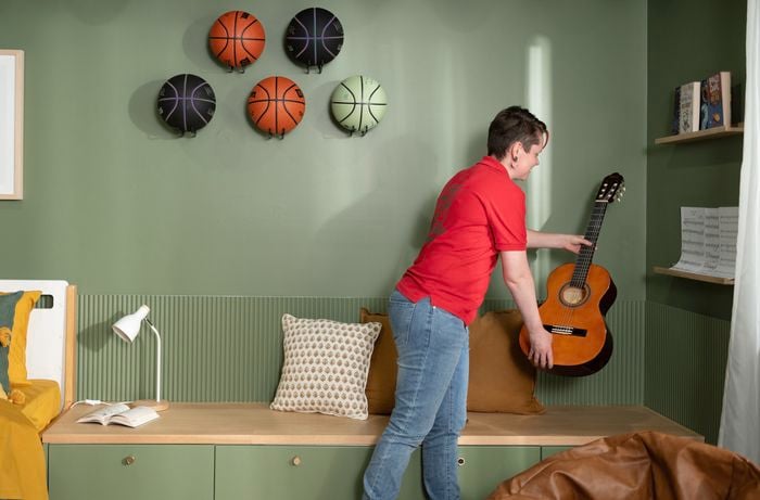 A Bunnings team member is holding a guitar in both hands in a neatly decorated room where five basketballs are hanging on the wall