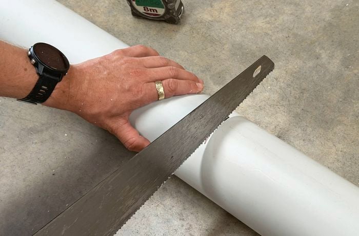 A hand saw is cutting through a white PVC pipe on a concrete floor.