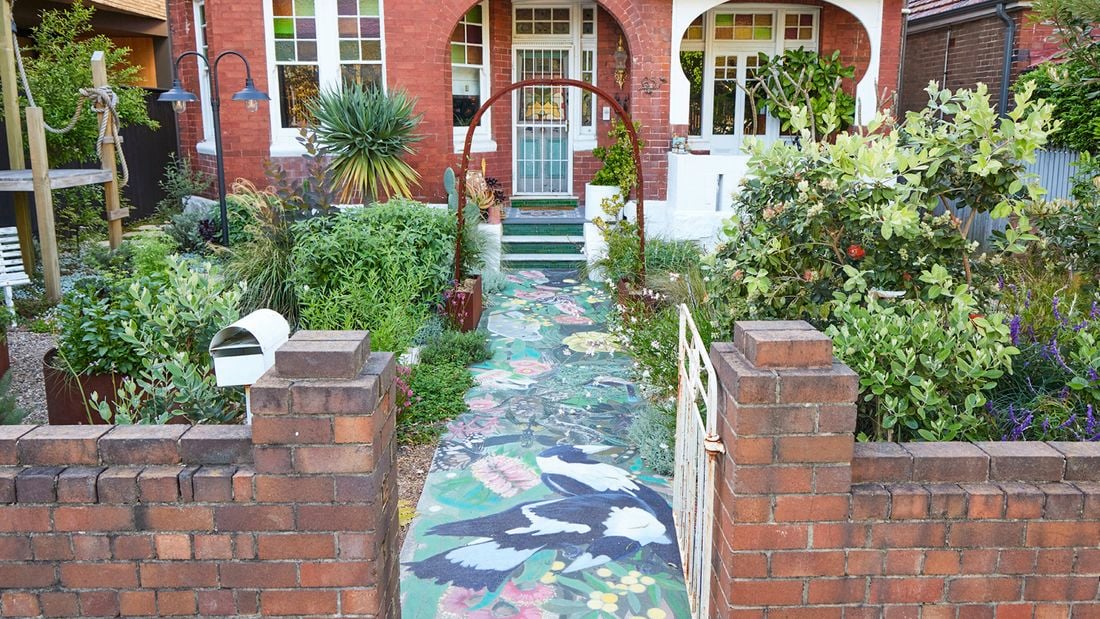 Front garden of a red‑brick period home with a white gate and a metal arch over a front path with a painted mural. Dense plants line the path, leading to a front porch with green tiled steps and a white security door.