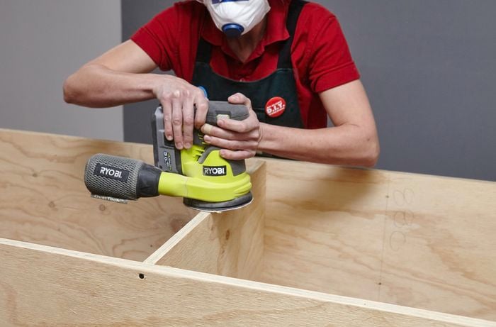 A person sanding the front end of a plywood shelf with an orbital sander