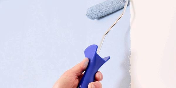 A hand holding a blue Monarch mini paint roller, painting pale blue paint onto a white wall.
