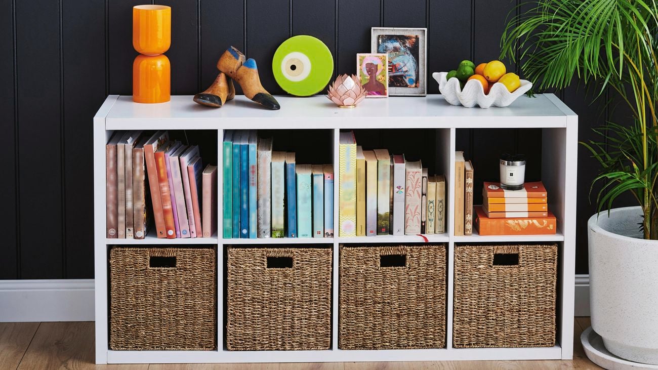 Clever cube shelving with woven inserts, including upright books and ornaments decorated on top of the unit with a bowl of fruit and a round mirror on dark blue backdrop.