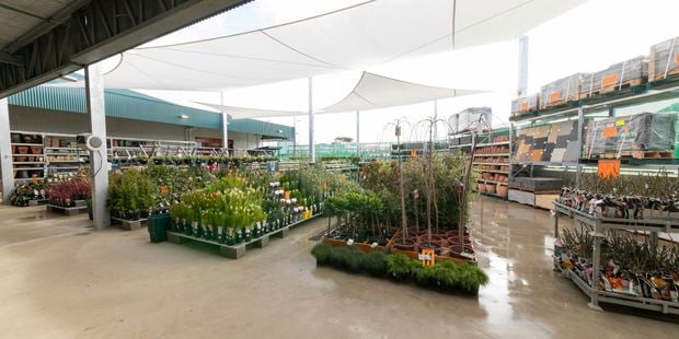 Open-air garden centre with rows of potted plants, shrubs and young trees displayed under large white shade sails. Wide concrete walkways run between plant displays, with shelving of pots and garden supplies visible along the perimeter of the covered outdoor retail space.