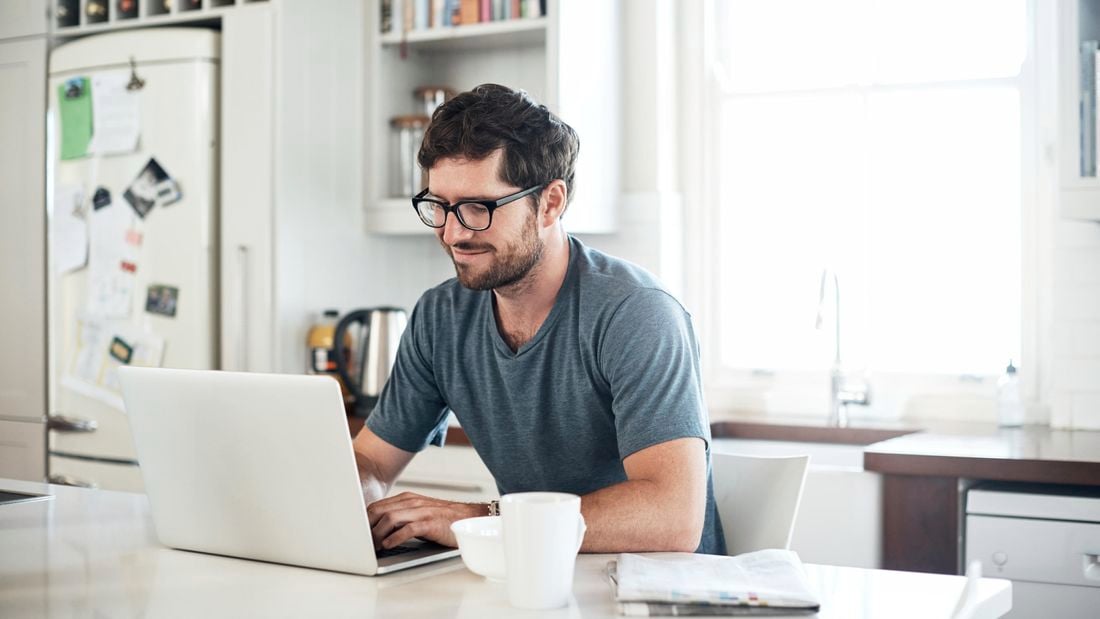 A smiling man using a laptop in his kitchen, with a cup of coffee within reach